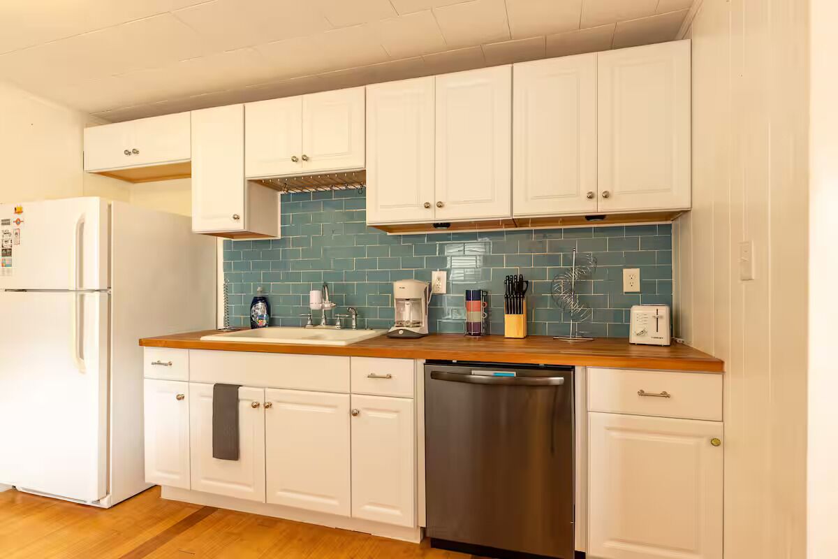 Cocina con armarios blancos, salpicadero de azulejos azules, lavavajillas de acero inoxidable y encimera de madera.