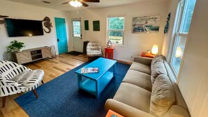 A living room with a blue coffee table, a beige sofa, a white chair, and a television mounted on the wall. - SandyPaws-0.5mi ToSound & 1mi ToBeach PetFriendly! (Kill Devil Hills)