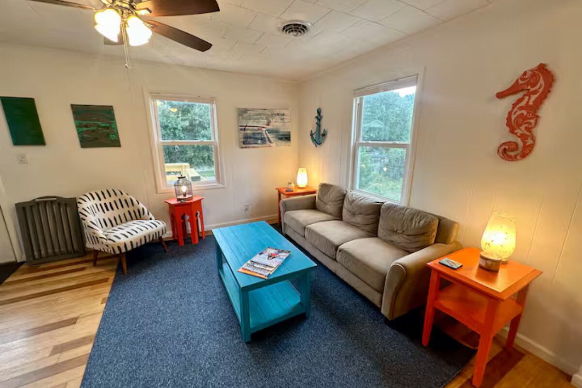 A living room with a blue coffee table, a beige sofa, a red side table with a lamp, a white armchair, and a ceiling fan.