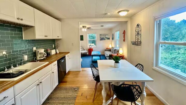A modern kitchen with white cabinets, a green backsplash, and a wooden countertop. There is a dining area with a white table and black chairs. - SandyPaws-0.5mi ToSound & 1mi ToBeach PetFriendly! (Kill Devil Hills)