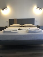 Cama bem arrumada com toalhas dobradas, cabeceira escura e duas luminárias fixadas na parede.