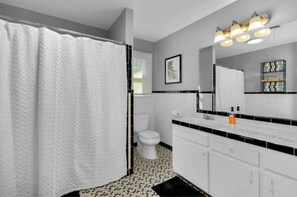 Ein Badezimmer mit Duschvorhang, Toilette, weißen Schränken, einem Waschbecken und einem beleuchteten Spiegel.