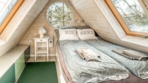 Een gezellig slaapkamer onder een schuin dak, een klein nachtkastje met lamp, een bed met twee kussens en uitzicht op bomen door een raam.