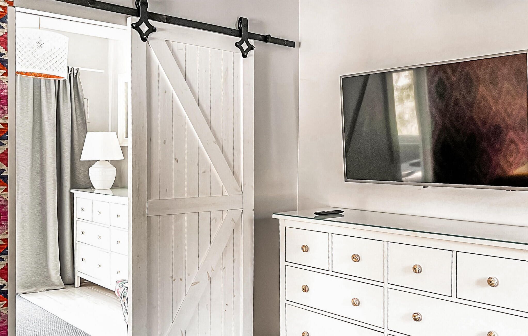 Una habitación con una puerta corredera de estilo rústico blanca, un aparador blanco con varios cajones, un televisor de pantalla plana montado en la pared y una lámpara sobre el aparador.