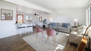 Un salon spacieux doté d’une grande fenêtre, un coin repas avec des chaises blanches, un canapé agrémenté de coussins décoratifs et une table basse.