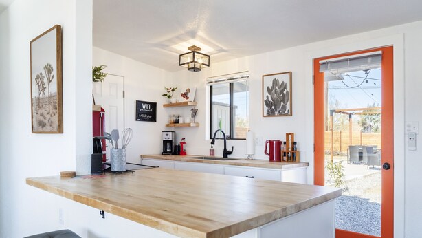 Une cuisine moderne avec un plan de travail en bois, des armoires blanches et une porte rouge donnant sur un patio.