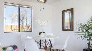Un coin repas avec une table ronde, des chaises blanches, un ventilateur de plafond et une grande fenêtre donnant sur des arbres et une clôture.