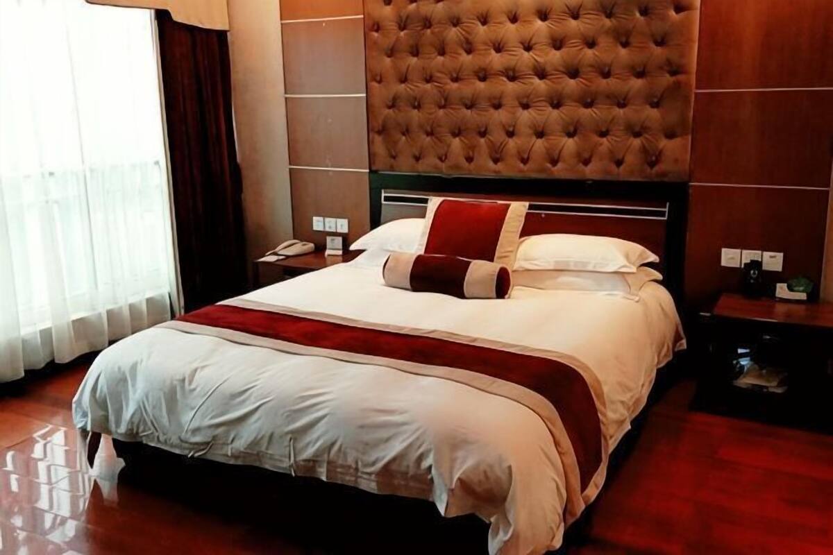 A hotel room with a large bed, a tufted headboard, a bedside table with a lamp, and a window with curtains.
