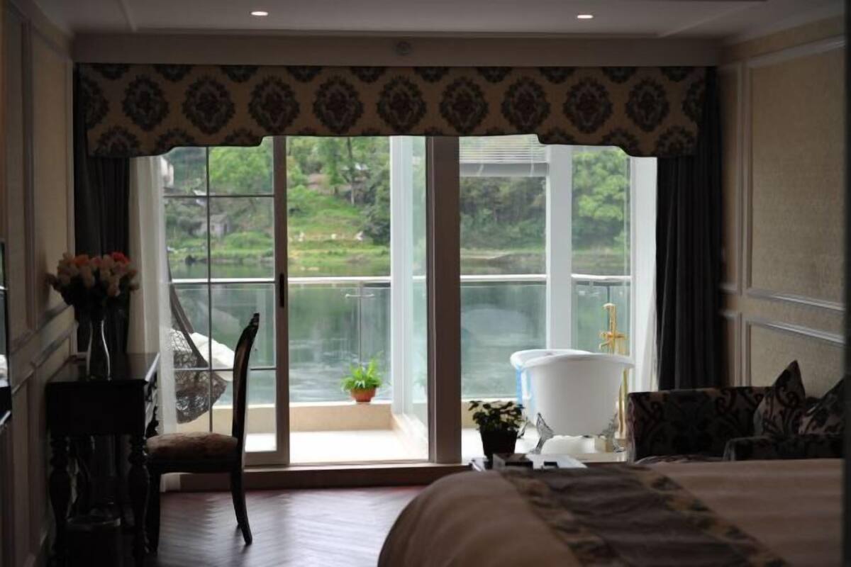 A bedroom with a large window, a bed, a chair, a small table, and a potted plant.
