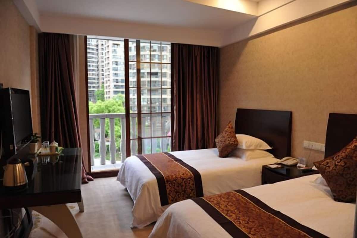 A hotel room with two beds, a desk, a TV, and a balcony with a view of high-rise buildings.