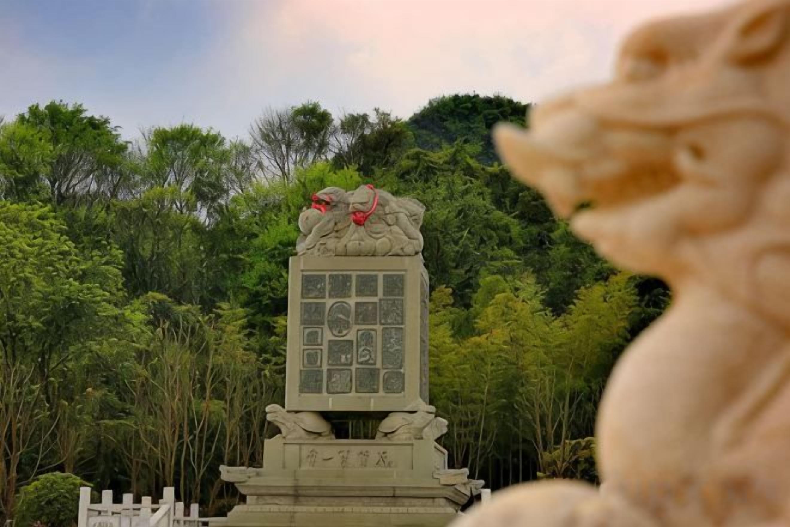 Un monumento in pietra con iscrizioni, circondato da un recinto bianco, sullo sfondo di alberi verdi e fitti.