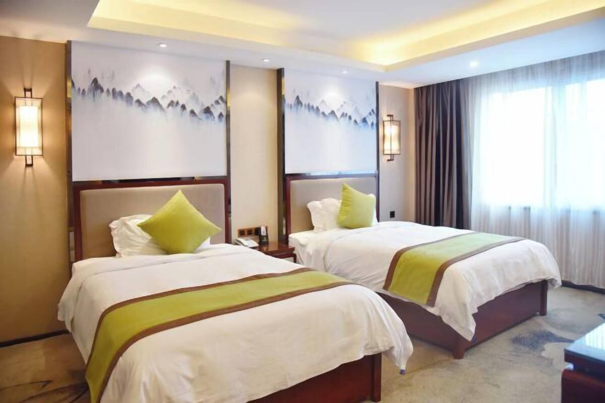 Une chambre d’hôtel avec deux lits, chacun recouvert d’une courtepointe blanche agrémentée de touches de vert, un grand tableau représentant des montagnes accroché au mur et une fenêtre avec des rideaux.
