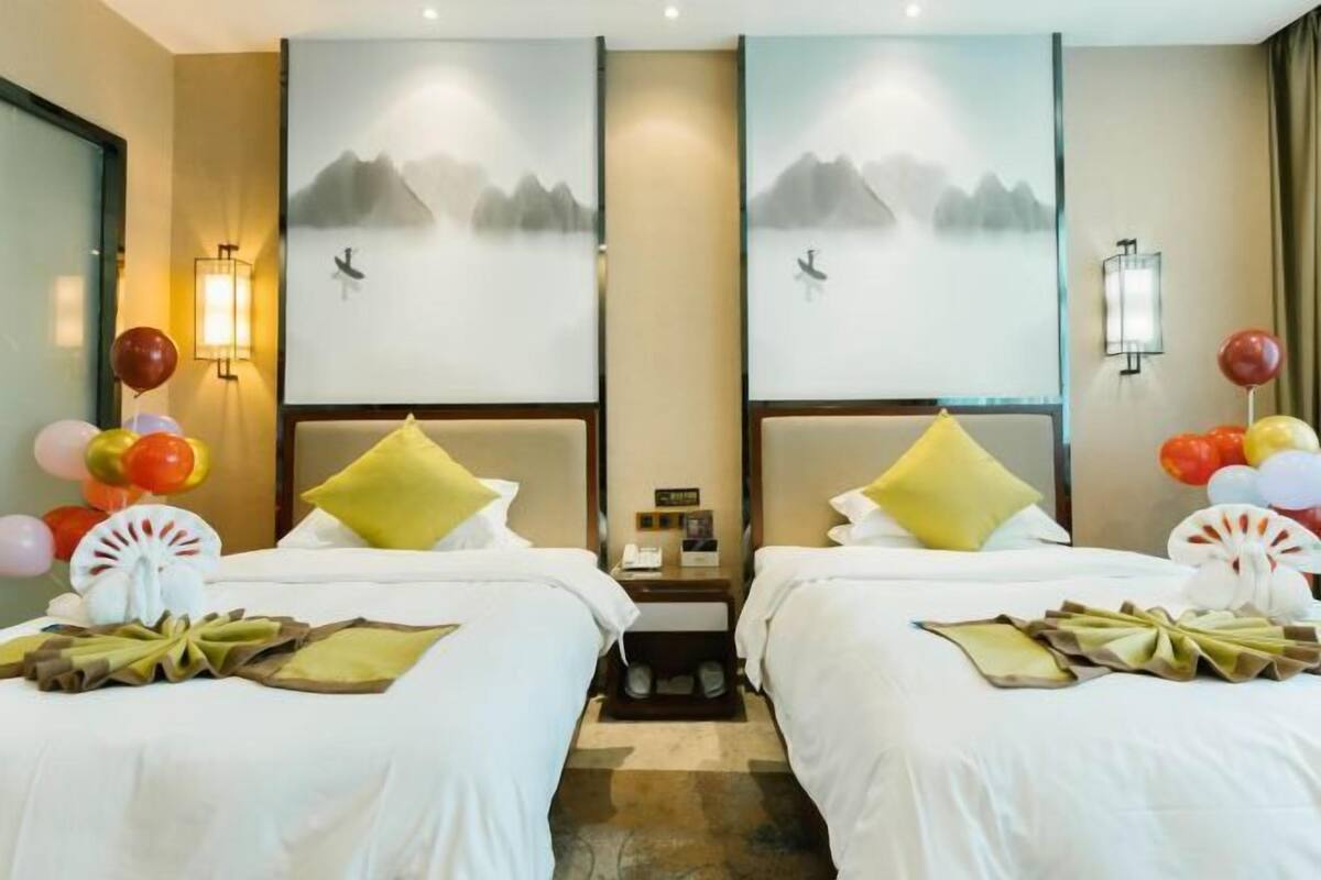 Une chambre d’hôtel avec deux lits, chacun étant équipé de draps blancs et d’oreillers jaunes. On y trouve des lampes fixées au mur et un tableau représentant des montagnes.