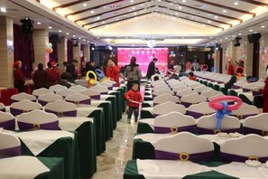 Ein Festsaal mit Tischen und Stühlen, eine Bühne mit rosafarbenem Hintergrund und Personen in roten Gewändern.