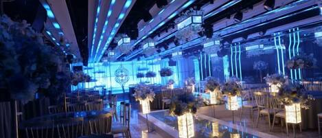 Un espacio con filas de mesas y sillas, un escenario con iluminación azul y centros de mesa florales.