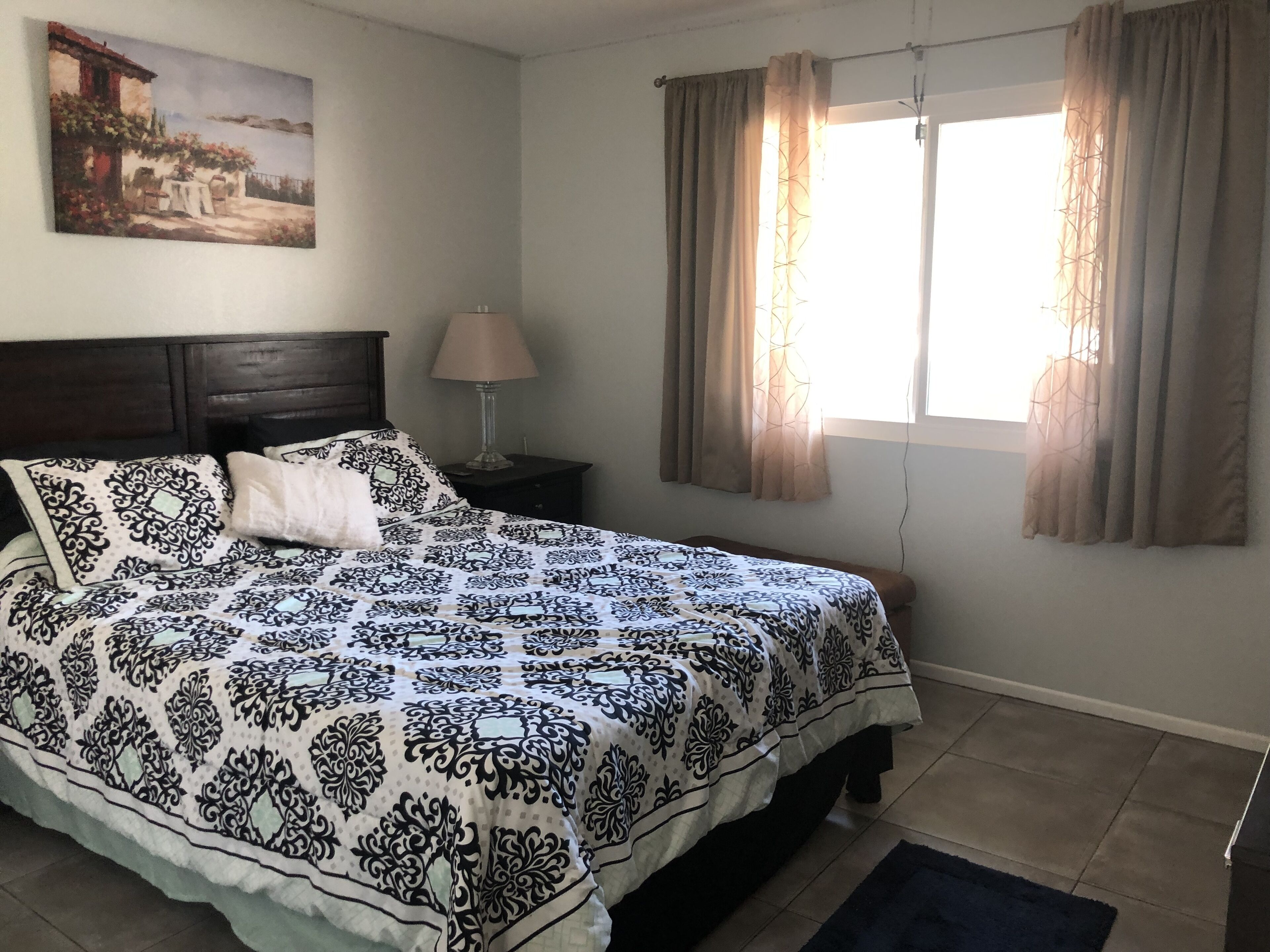 A bedroom with a bed, a nightstand, a lamp, a window with curtains, and a painting on the wall.