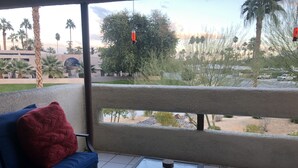 Un balcón con un sofá azul, una almohada roja, una mesa de vidrio con una planta y vasos, y vistas a palmeras y un edificio.