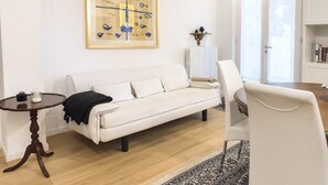 Un salon avec un canapé blanc, une table ronde en bois, un fauteuil blanc, un tapis à motifs, une œuvre d’art encadrée accrochée au mur et un radiateur.