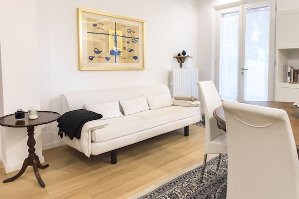 A living room with a white sofa, a round wooden table, a white armchair, a patterned rug, a framed artwork on the wall, and a radiator.