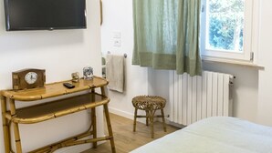 Une chambre avec un lit, un téléviseur fixé au mur, une petite table avec une horloge et une télécommande, un tabouret en osier et une fenêtre avec des rideaux.