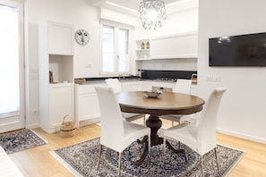 A modern kitchen with white cabinets, a dining table with chairs, a chandelier, and a flat-screen TV.