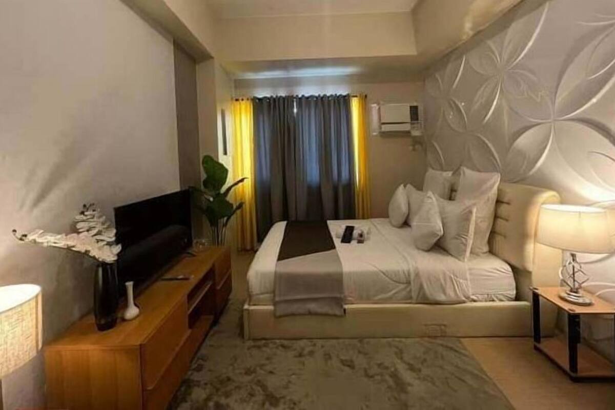 A hotel room with a bed, a TV on a wooden stand, a bedside table with a lamp, and a decorative plant.