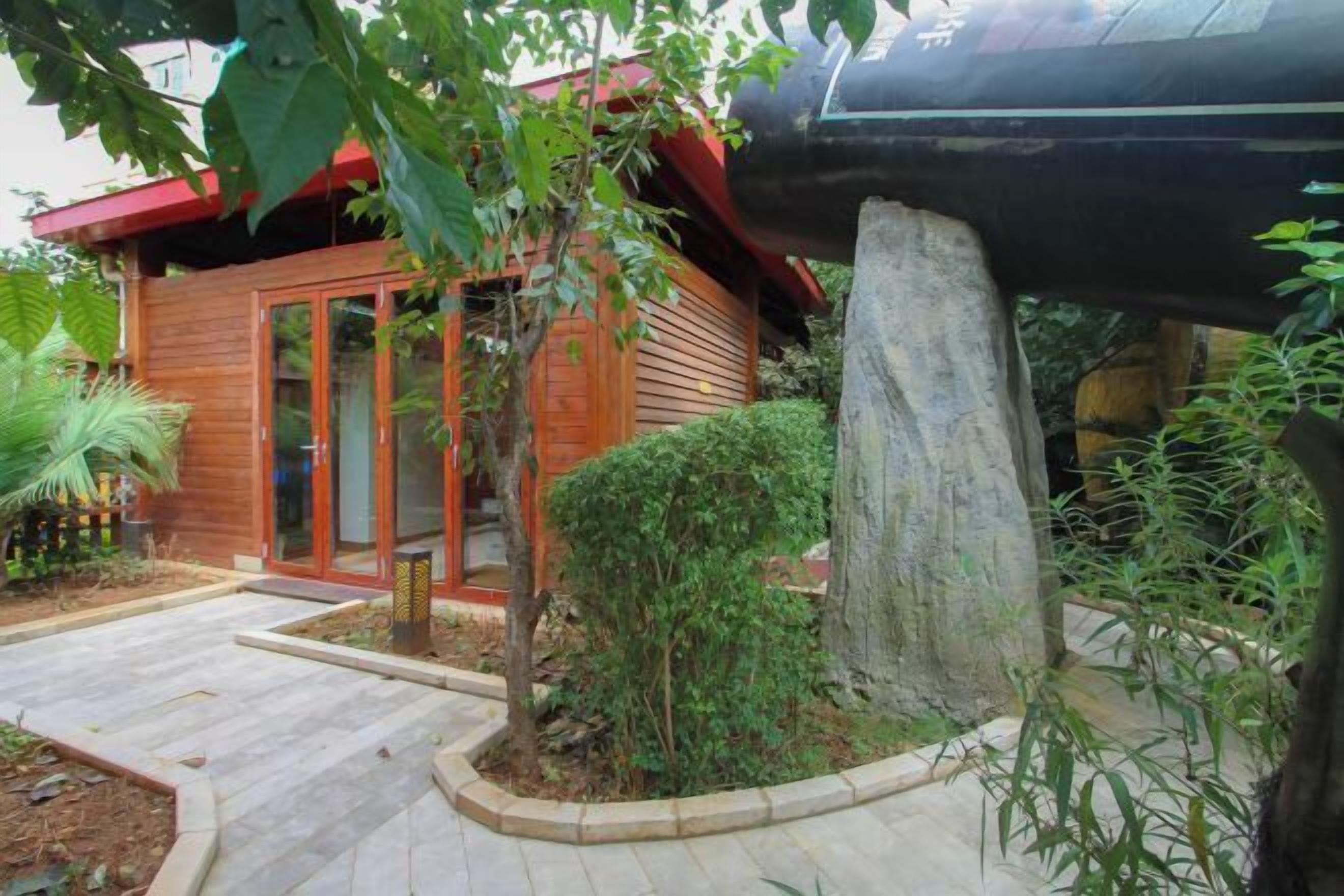 Uma casa de madeira com porta de vidro, rodeada por um jardim com um pilar de pedra e diversas plantas.