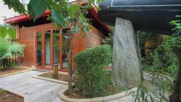 Uma casa de madeira com porta de vidro, rodeada por um jardim com um pilar de pedra e diversas plantas.