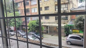 Visão pela janela com grades de metal, mostrando carros estacionados, uma rua e edifícios ao fundo.