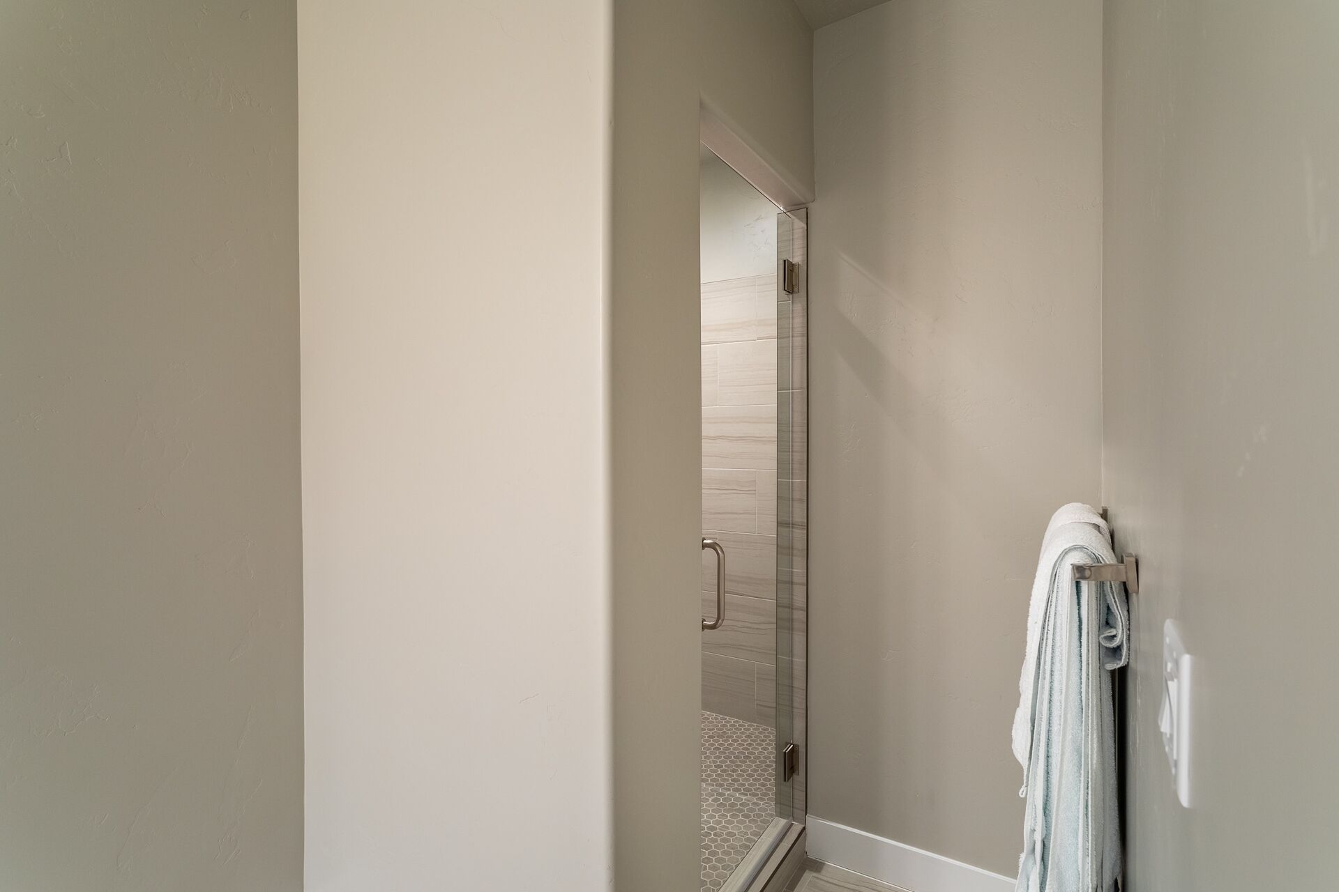 Un couloir menant à une salle de bain dotée d’une porte de douche en verre et d’un porte-serviettes avec une serviette.