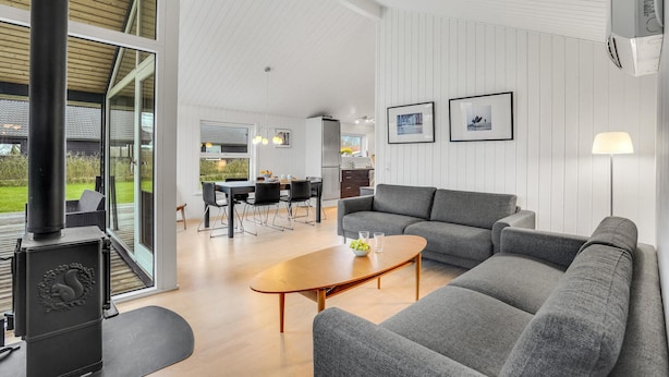 A modern living room with a grey sofa, a wooden coffee table, and a black stove. There is a dining area with a table and chairs, and a kitchen in the background.