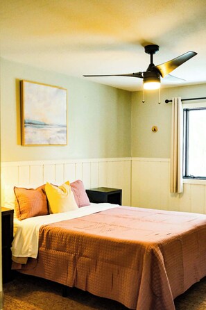 A bedroom with a bed, a ceiling fan, a bedside table, a nightstand, a window, and a painting on the wall. - Winter Ski Retreat at Powderhorn Mountain (Ironwood Township)