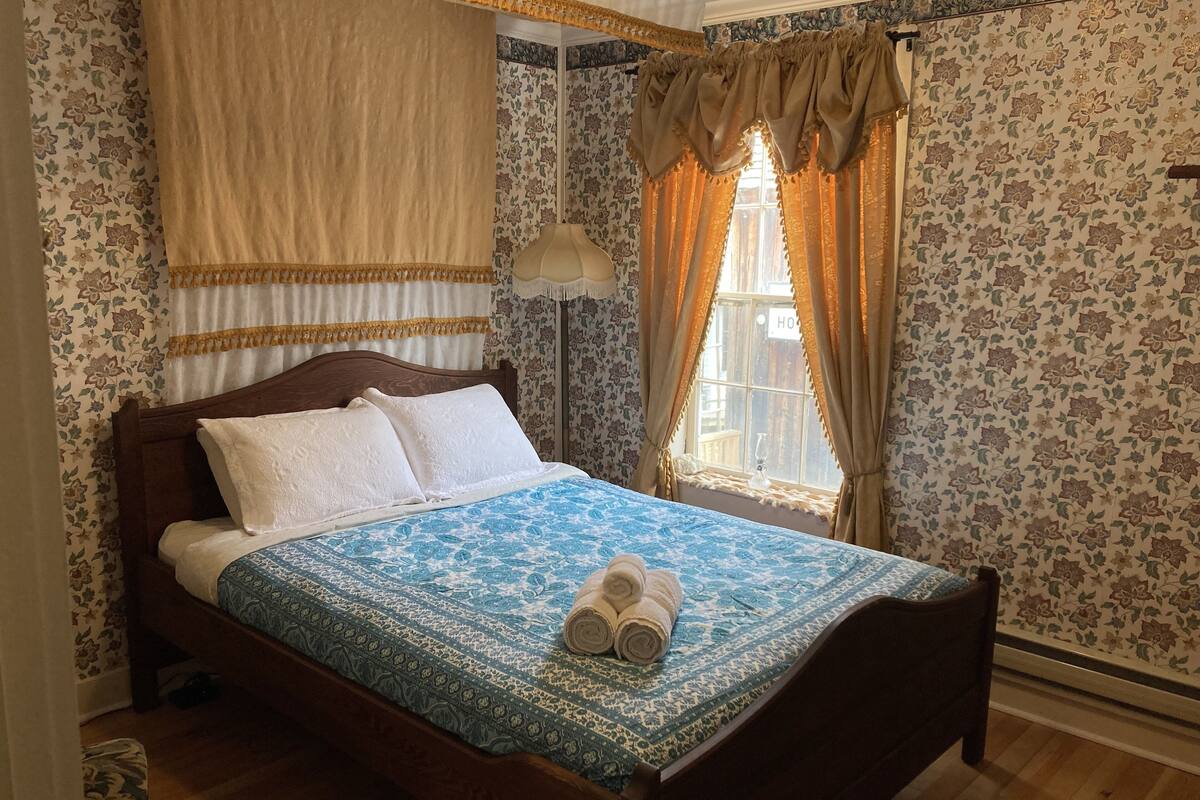 Un lit bien fait, recouvert d’une couvre-lit bleue à motifs, avec une tête de lit en bois et deux serviettes enroulées au pied du lit.