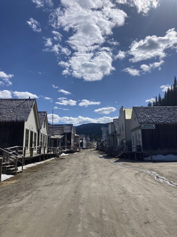 A historic town street with buildings, a dirt road, and a mountainous backdrop.