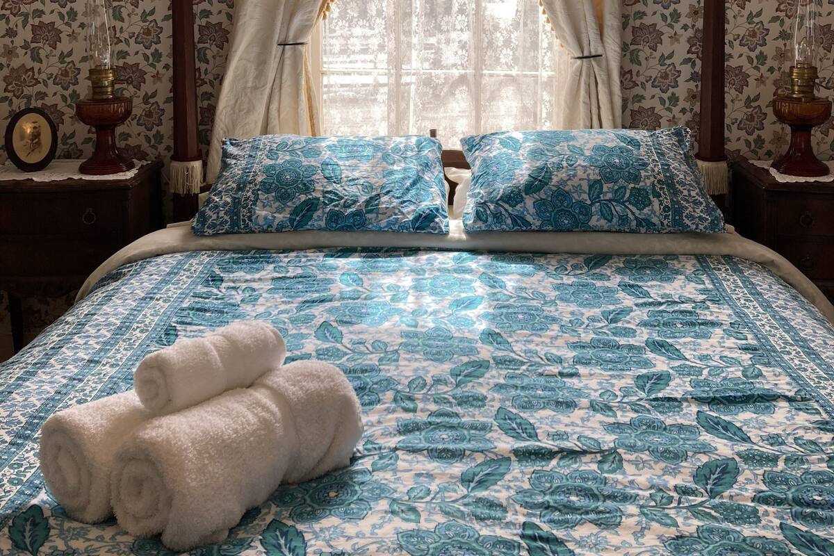 Une chambre à coucher avec un papier peint à motifs floraux, un lit avec une couette bleue et blanche, deux oreillers, une fenêtre avec des rideaux et une table de chevet avec une lampe.