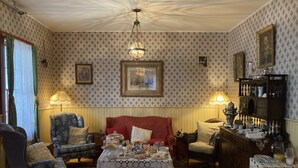 A cozy room with patterned wallpaper, a red sofa, a blue armchair, a table with various items, and a chandelier. - St. George Hotel (Barkerville)