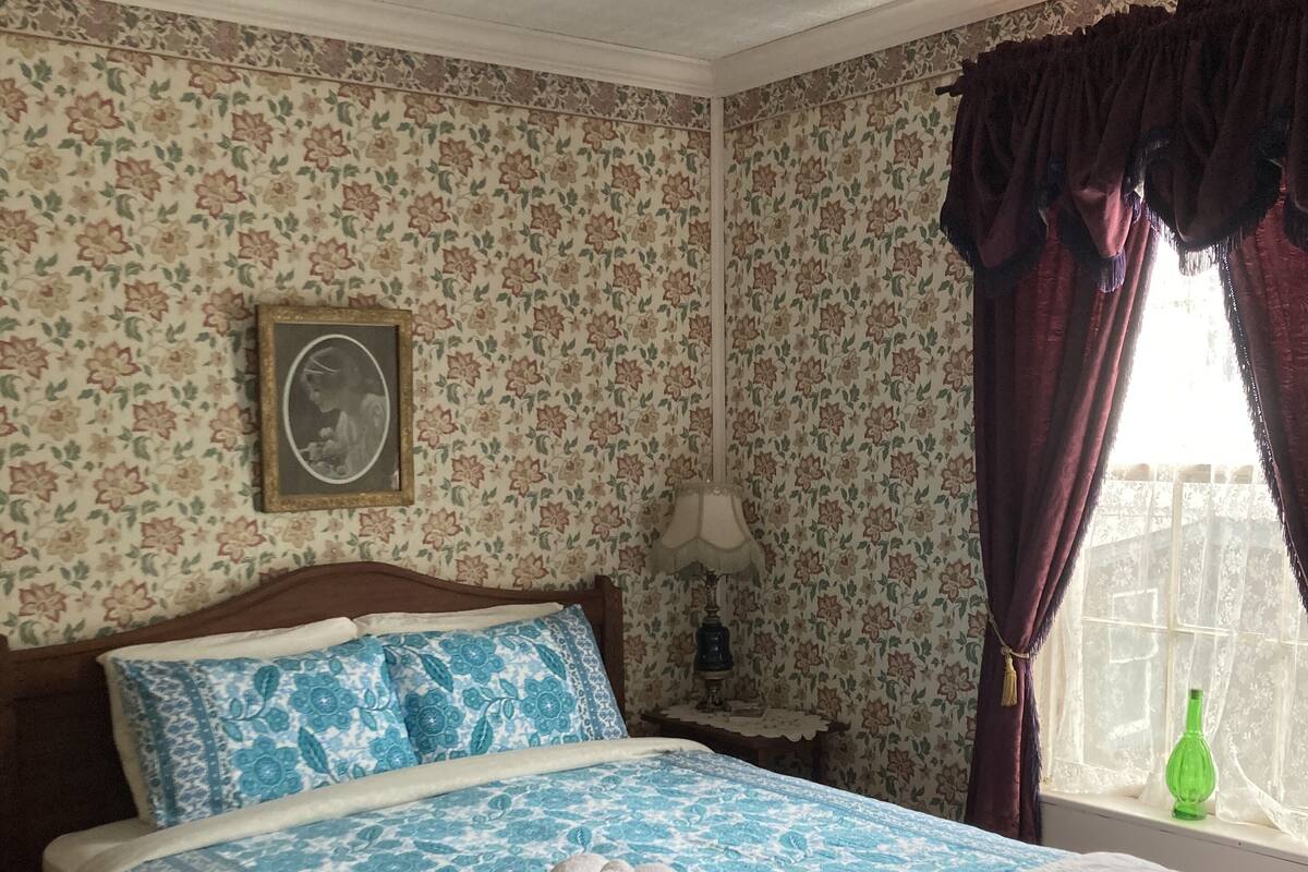 Une chambre avec un papier peint à motifs floraux, un lit en bois avec une literie bleue et blanche, une table de chevet avec une lampe et une fenêtre avec des rideaux.