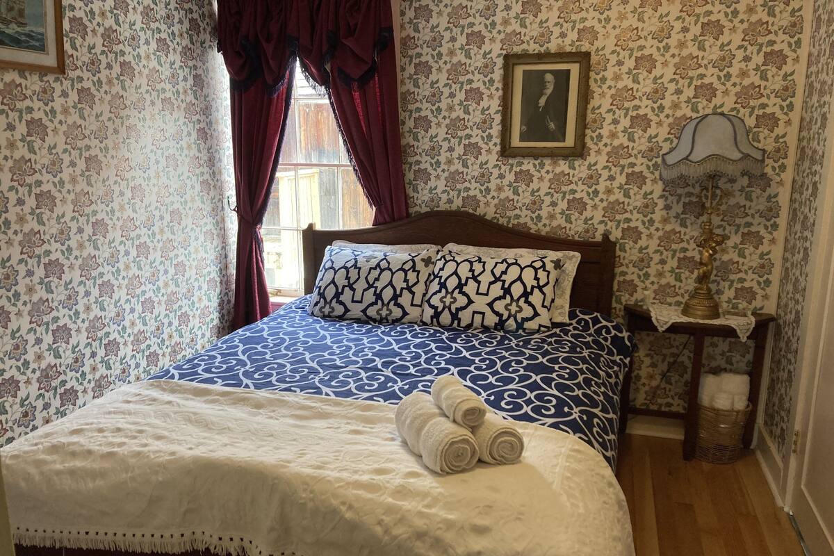 Un lit bien fait, recouvert d’une courtepointe bleue et blanche à motifs, d’un plaid blanc et agrémenté de serviettes enroulées au pied du lit.