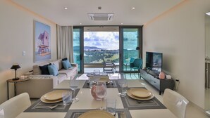 A modern living room with a dining area, large windows, a flat-screen TV, and a view of the countryside. - A-1402 STUNNING 1 BEDROOM (Cupecoy)
