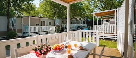 A covered outdoor dining area with a table set for breakfast, surrounded by white railings and a view of mobile homes.