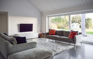 A modern living room with a grey sofa, a flat-screen TV mounted on the wall, and a large window providing a view of the outdoors. - Beach front home in Skivarp with house sea view (Skivarp)