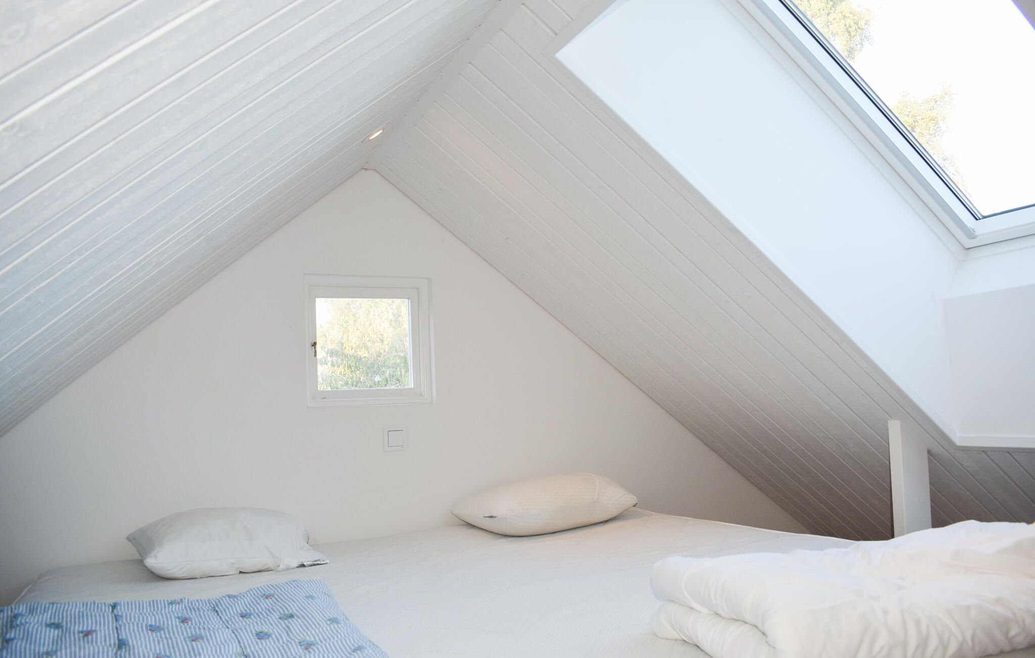Una habitación pequeña con techo inclinado, paredes blancas, una ventana, una cama con almohadas y una manta azul a rayas.
