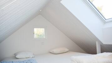 Una habitación pequeña con techo inclinado, paredes blancas, una ventana, una cama con almohadas y una manta azul a rayas.
