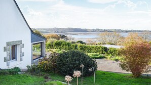 A white house with a stone facade, a garden with various plants, and a view of a body of water and distant landscape.