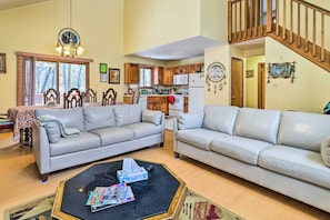 A spacious living room with two light-colored sofas, a coffee table, and a dining area with wooden cabinets and a chandelier. A staircase leads to an upper level.