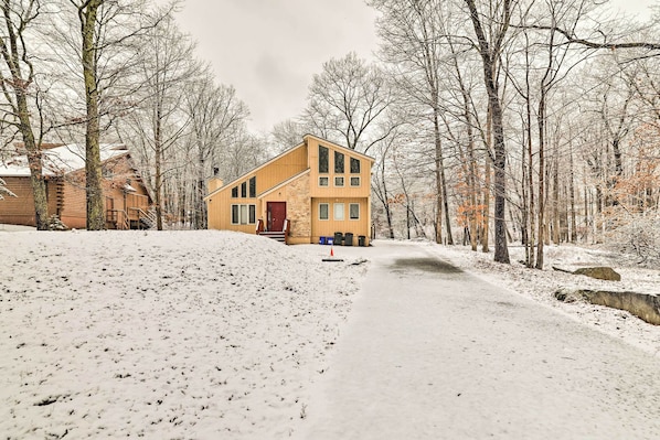 A two-story house with a red door and multiple windows, surrounded by snow-covered ground and leafless trees.