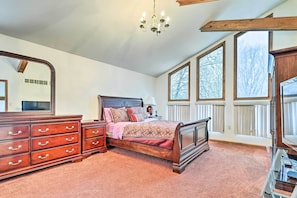A bedroom with a large bed, wooden dresser, and a mirror. There are large windows with blinds, a chandelier, and a radiator.
