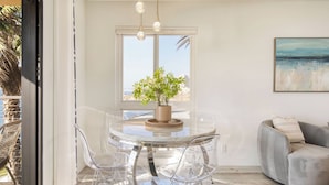 A modern dining area with a glass table, clear chairs, a potted plant, and a view of the sea through a large window. - Oceanfront in La Jolla, Steps to Beach, Shops, Dining Dawn Patrol by Avantstay (La Jolla)