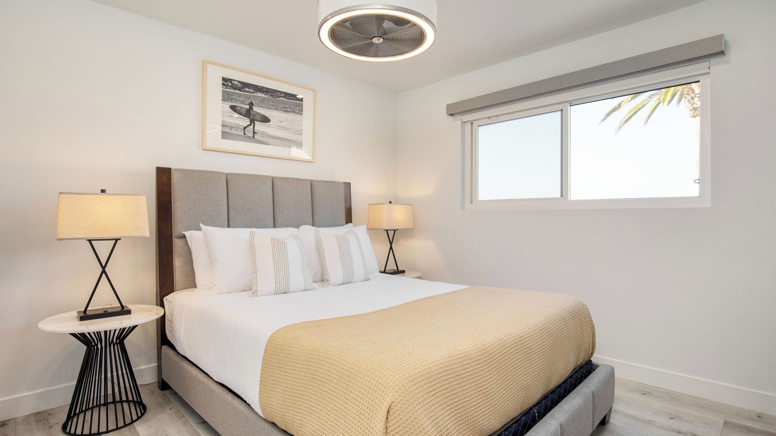 Une chambre à coucher bien rangée, avec un lit, deux tables de chevet, un ventilateur de plafond, une fenêtre avec des stores et un tableau encadré au mur.