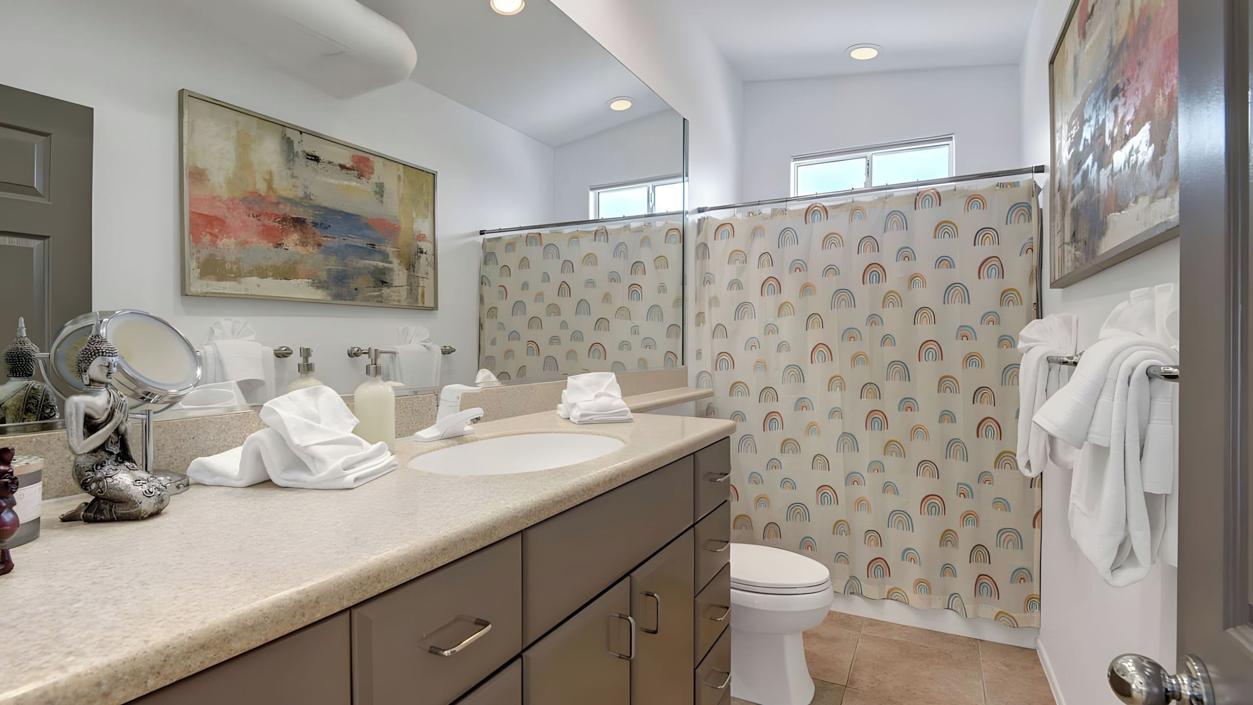 A modern bathroom with a large mirror, two sinks, a toilet, and a shower with a rainbow pattern.